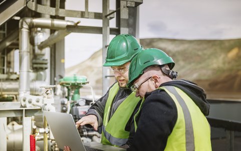 Two safety workers in hard hats, safety glasses and high-visibility vests reviewing data on a laptop at an industrial plant